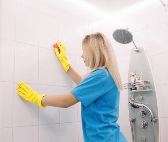 Woman with special cleaning sponge cleaning wall tiled surface in bathroom. Blonde woman, employee cleaning company, wearing in yellow protective rubber gloves, with special cleaning sponge, cleaning wall tiled surface in white bathroom. Housework and housekeeping concept.